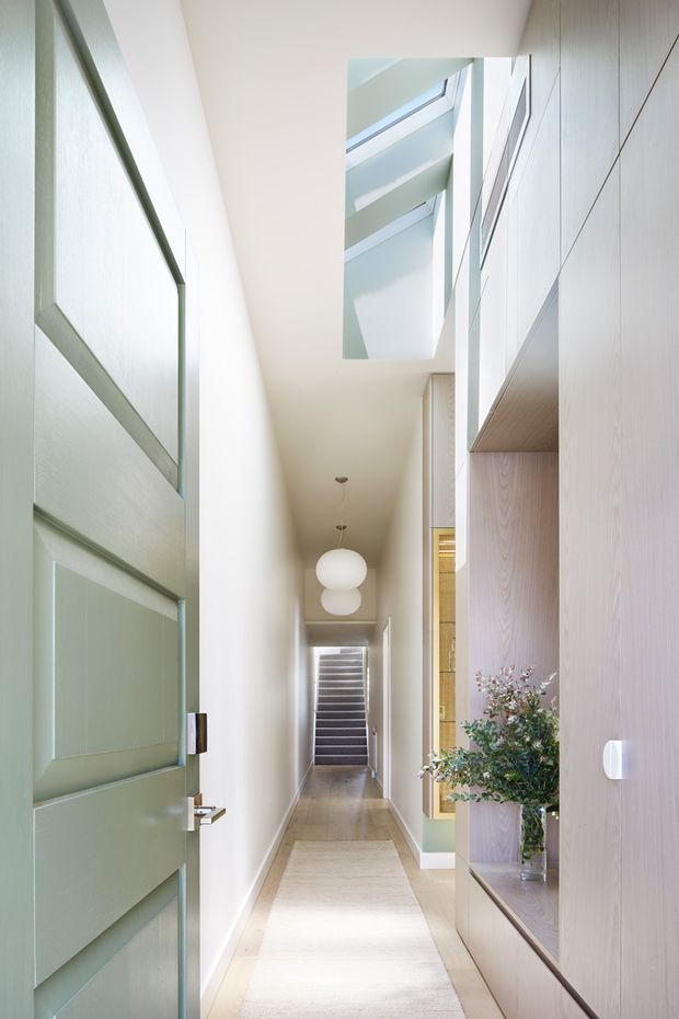 A bright, narrow residential hallway viewed from an open pale green paneled front door. The corridor has light walls, a timber-toned floor, a long neutral runner, spherical pendant lights, and built-in pale timber cabinetry with open display shelving on the right. A stairway is visible at the far end, while a skylight or clerestory opening above brings in abundant natural light.