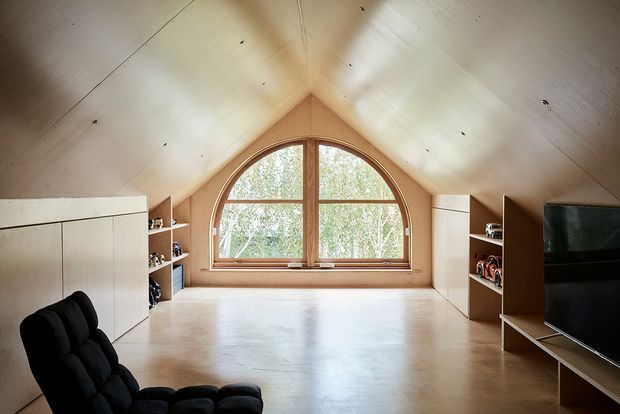 A warm attic-style media or living room with a steeply pitched, timber-lined ceiling and a large arched timber-framed window at the gable end. Built-in white storage cabinetry and open shelving line both sides, with a television on the right and a black lounge chair in the foreground. The room has a bright, minimal, contemporary feel with pale timber surfaces and a smooth light floor.