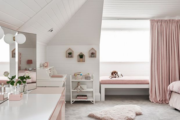 Bright, softly styled bedroom or children's room with white painted shiplap walls and ceiling, a large window seat upholstered in blush pink, floor-length blush curtains, white built-in cabinetry and shelving, and a pale plush rug. A bed is partially visible at right, while a mirrored vanity or dresser occupies the left foreground. The room has a pitched ceiling and abundant natural light.