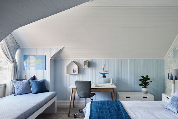 Bright, compact coastal-style bedroom with a built-in window seat, twin beds, blue bedding and cushions, and a small study desk against pale blue vertical wall panelling. The room has a distinctive white curved and panelled ceiling, soft natural light, nautical decor, and a calm blue-and-white palette.