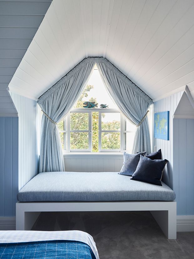 A bright, compact coastal-style bedroom nook with a built-in window seat beneath a steeply pitched, white panelled ceiling. Soft blue-grey gathered curtains frame the central multi-pane window, while blue cushions and pale blue upholstery create a calm palette. A partial bed with blue checked bedding is visible in the foreground, and blue-and-white wall panelling surrounds the space.