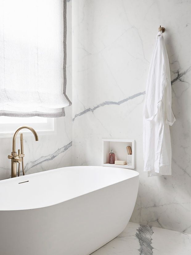 A bright, refined bathroom featuring a sculptural white freestanding bathtub, white marble wall and floor surfaces with prominent grey veining, and a brushed brass wall-mounted bath filler. A softly filtered window with a white Roman blind provides natural light. A recessed wall niche holds toiletries, while a white robe hangs from a small brass hook.
