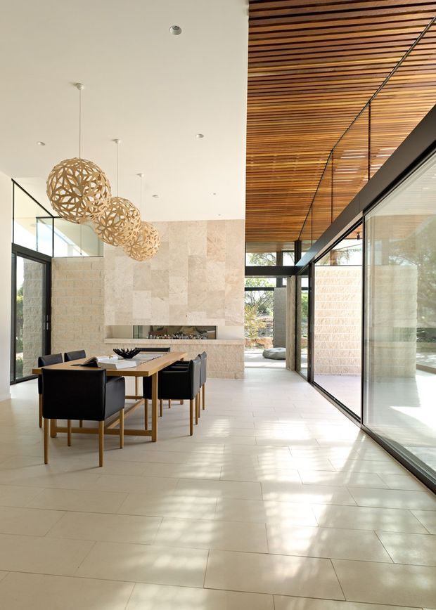 Bright, refined open-plan dining area with a long timber dining table and dark upholstered chairs. The space features pale stone wall cladding around a linear fireplace, large black-framed floor-to-ceiling glazing, a warm slatted timber ceiling, pale tiled flooring, and sculptural woven pendant lights. The glazing connects the dining space to a light-filled courtyard or garden beyond.