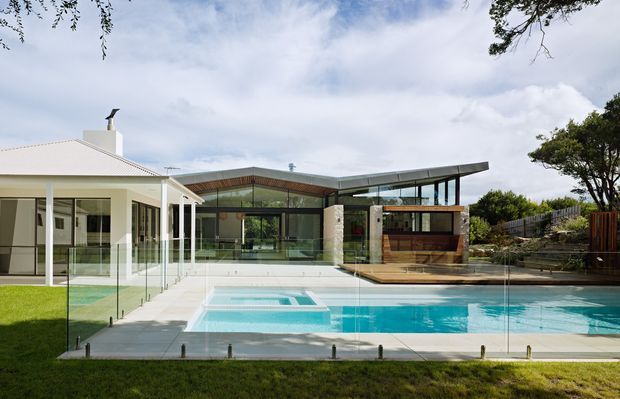 Exterior view of a contemporary residential property with a large swimming pool in the foreground. The composition shows a white pitched-roof house connected to a modern flat-roof pavilion with extensive floor-to-ceiling glazing, timber screening, and pale stone walls. A frameless glass pool barrier separates the lawn from the pool terrace, with landscaped garden areas around the building.