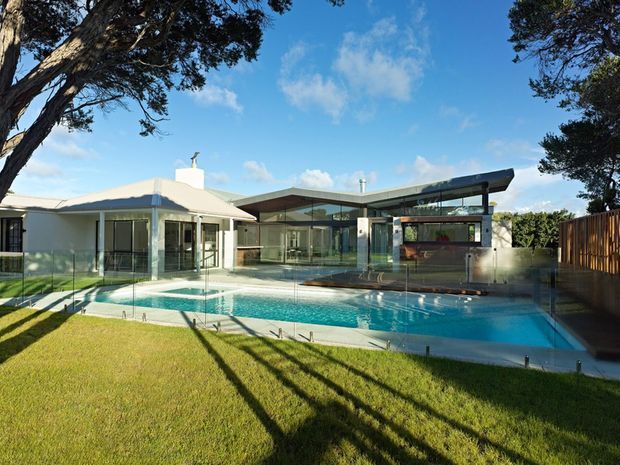 A sunny exterior view of a contemporary residential property centred around a large swimming pool. The home combines a light-coloured low-pitched roofed wing with a modern pavilion featuring extensive floor-to-ceiling glazing, dark structural framing, and a broad projecting roof. Glass pool fencing, timber screening, paved pool surrounds, and landscaped lawn are visible.
