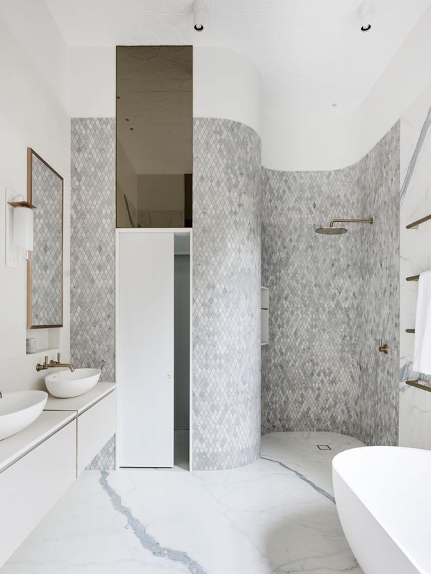 Bright contemporary bathroom with a sculptural curved shower enclosure clad in small grey diamond-patterned mosaic tiles. The room features a large white freestanding bathtub, double white vessel basins on a streamlined vanity, a tall mirrored cabinet, brass-toned shower and tap fittings, and white marble-look surfaces with pronounced grey veining. White curved ceiling forms and recessed lighting create a clean, refined appearance.