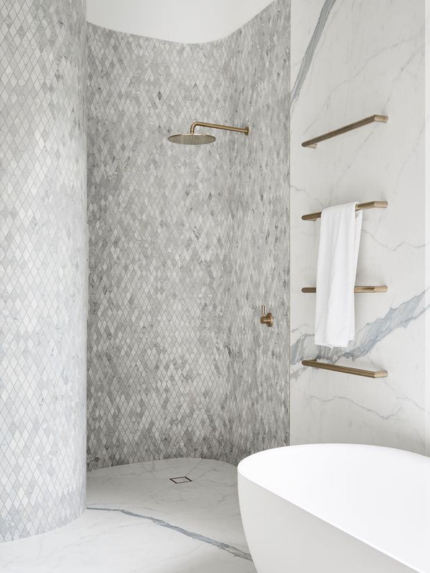 A refined contemporary bathroom featuring a curved walk-in shower enclosure lined with small grey diamond-patterned mosaic tiles. Large white marble-effect wall and floor surfaces with grey veining contrast with brushed brass shower and towel hardware. A freestanding white bathtub is partially visible in the foreground. The palette is light, monochromatic, and luxurious.