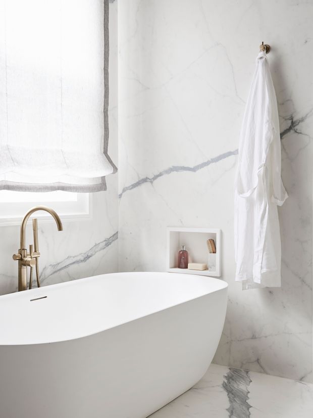 Bright, luxurious bathroom featuring a freestanding white oval bathtub, full-height white marble-look wall and floor surfaces with prominent grey veining, a brass bath filler, recessed wall niche, roller blind, and white robe hanging from a brass hook. The space has a clean, refined, spa-like appearance with abundant natural light.