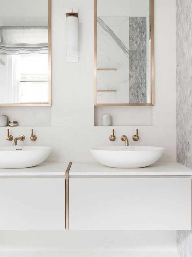 Bright, refined bathroom vanity with two white vessel basins set on a floating white cabinet. Brushed brass wall-mounted taps and slim mirror frames add warmth, while large marble-look wall surfaces and a patterned vertical tile panel create subtle contrast. The mirrors reflect a window and shower or wall finishes.
