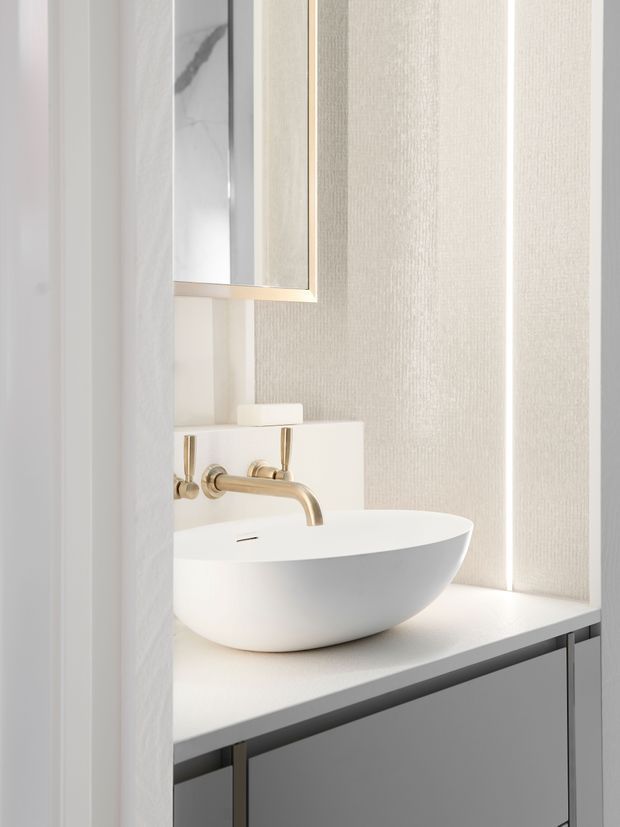 Close-up view of a refined bathroom vanity with a white vessel basin, brushed brass wall-mounted tapware, pale stone or solid-surface benchtop, grey built-in cabinetry, textured light wall finish, and a large framed mirror. The space is bright, minimal, and softly neutral with warm metallic detailing.