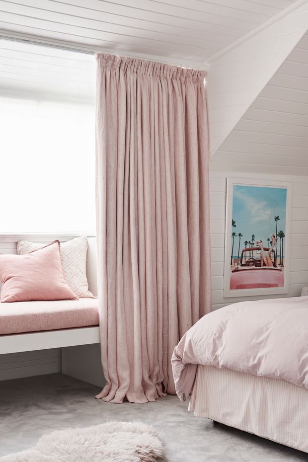 A bright, soft-toned bedroom with white painted shiplap walls and ceiling, a sloped ceiling detail, blush pink floor-length curtains, a built-in window seat with pink cushions, and a bed dressed in pale pink bedding. The room has grey carpet, a fluffy pale rug, and a framed pastel artwork, creating a gentle, playful atmosphere.