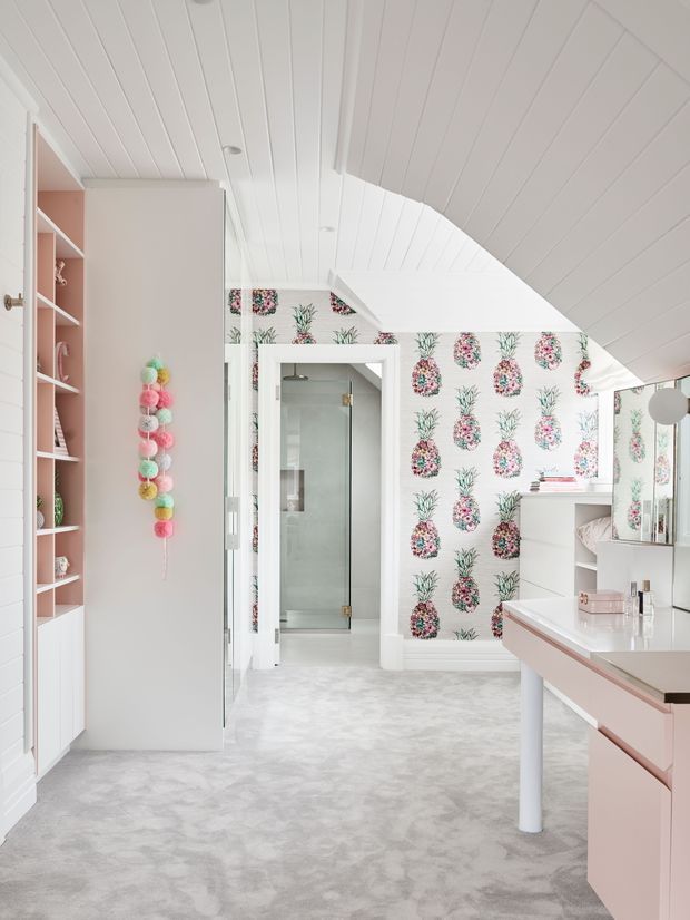 Bright attic-style bedroom dressing area with a sloped white panelled ceiling, pale pink built-in shelving and vanity cabinetry, soft grey carpet, and a glass-enclosed bathroom visible through the doorway. A bold pineapple-patterned feature wallpaper adds a playful tropical accent, while mirrored cabinetry and pastel decorative details reinforce the feminine, youthful styling.