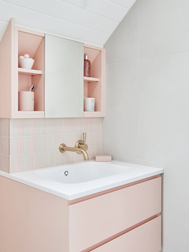 A compact, softly styled bathroom vanity with blush-pink cabinetry and open shelving, a white integrated basin, brushed brass wall-mounted tapware, and a vertically ribbed pale pink tile splashback. Large light grey-white wall tiles and a white panelled ceiling create a clean, bright, pastel-toned interior.