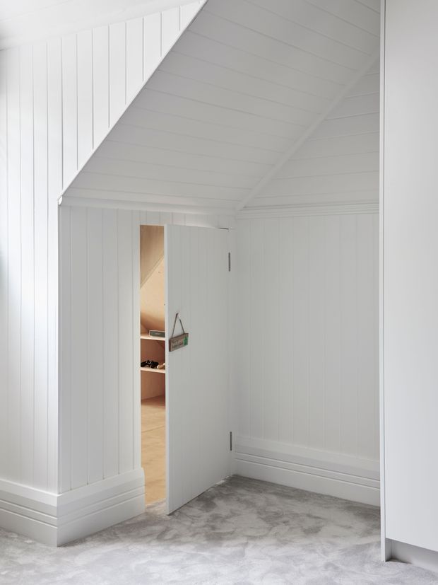 A small, bright white-lined interior space with vertical wall panelling, a sloped panelled ceiling, and white decorative skirting. A partially open door leads into a warmer timber-finished room, while the main space has a grey patterned carpet and minimal furnishings.
