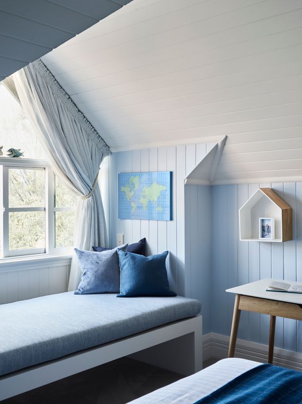 A bright, compact coastal-style bedroom or children’s room with pale blue vertical wall panelling, a sloped white-lined ceiling, and a daybed beneath a window. Blue cushions, layered blue-and-white textiles, soft drapery, a small wall-mounted world map, and a simple wood-legged desk create a calm nautical feel. A house-shaped wall shelf provides playful storage.
