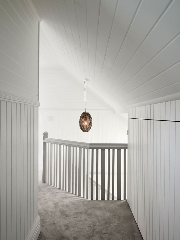 Bright white upper-level hallway and stair landing with vertical wall panelling, a sloped paneled ceiling, white balustrades, and grey carpet. A woven pendant light hangs over the stair opening, creating a warm focal point in the otherwise light interior.