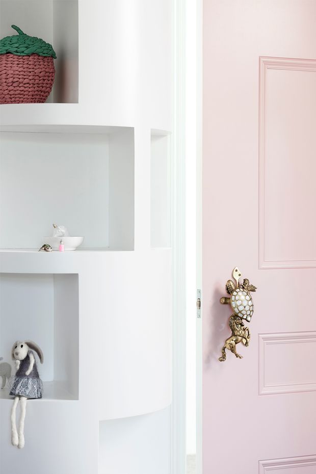 A bright, playful interior detail showing white built-in recessed shelving beside a pale pink raised-panel door. The shelves contain small decorative objects and toys, including a knitted fruit-shaped basket and a fabric doll. The door has ornate brass-gold animal-themed hardware, creating a whimsical children's-room aesthetic.
