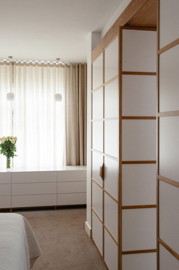 A bright, refined bedroom featuring a large white built-in wardrobe with warm timber framing and panelled sliding doors. A low white dresser sits beneath sheer and beige curtains, with soft pendant lights, a vase of flowers, and part of a neatly made bed visible. The room has a calm neutral palette and carpeted floor.