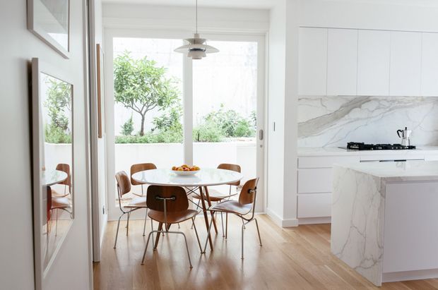 Bright open-plan kitchen and dining area with white flat-panel cabinetry, a veined stone backsplash and island, pale timber flooring, and a round white dining table surrounded by mid-century style wooden chairs. Large glazed doors open toward a planted outdoor area, while a simple pendant light and framed artwork add understated detail.