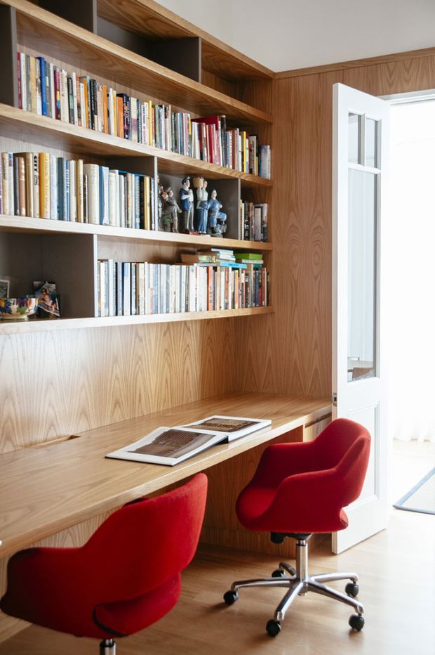 A compact home office lined with warm timber wall panelling and custom open bookshelves filled with books and decorative objects. A long built-in timber desk spans the wall, paired with two vivid red upholstered swivel chairs. A white glazed door opens to a bright adjacent space.