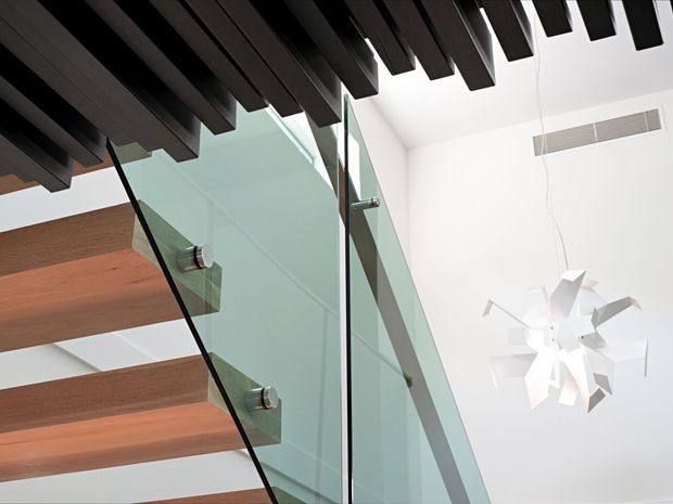 Architectural interior detail looking upward at a contemporary stairway. Thick timber stair treads are supported beside a clear glass balustrade with metal fixings, while dark exposed ceiling battens create a strong linear pattern overhead. A sculptural white pendant light hangs in the bright double-height space, with a wall-mounted ventilation grille visible in the background.