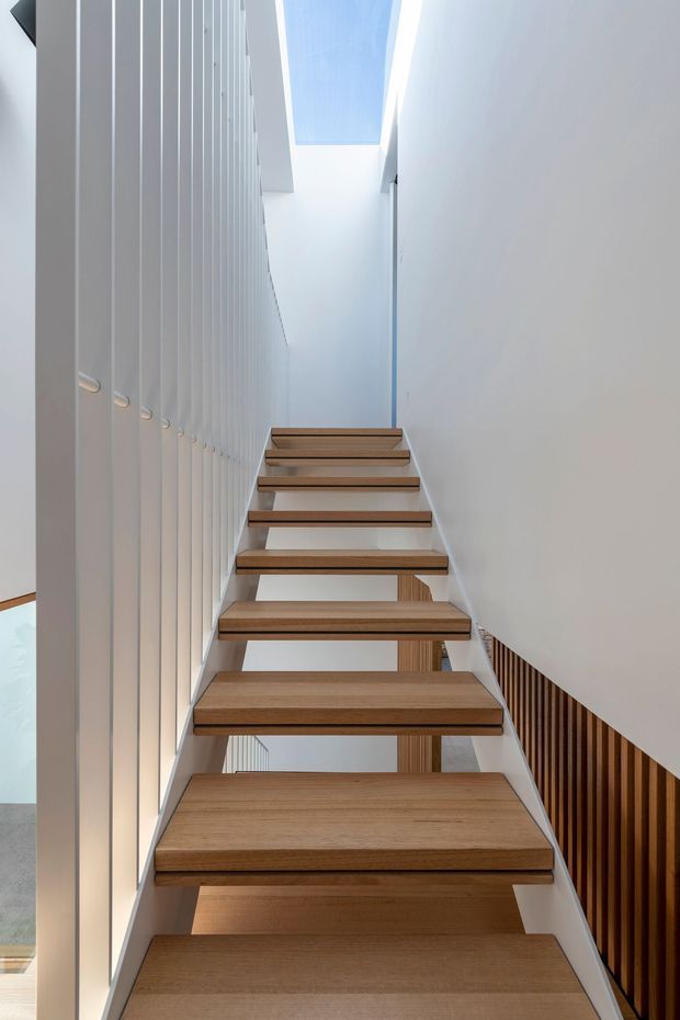 A minimalist interior staircase viewed from below, with floating warm timber treads, slim pale structural supports, a tall white vertical-panel wall on the left, and a dark timber slatted balustrade or screen on the right. A narrow skylight or clerestory above brings in bright natural light, creating a clean, airy vertical composition.