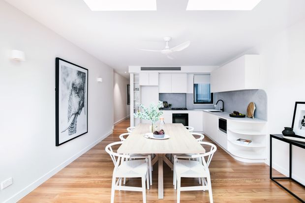 Bright open-plan dining and kitchen interior with a light timber dining table surrounded by white chairs, white contemporary cabinetry, a curved grey stone-look benchtop and splashback, timber flooring, recessed ceiling lighting, and a ceiling fan. A framed monochrome artwork and slim black console add contrast.