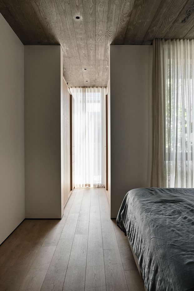 A calm, minimalist bedroom with a partially visible bed in the foreground, tall sheer-curtained glazing, pale timber flooring, and a distinctive continuous timber-lined ceiling extending down the central passage. Built-in wardrobes or wall volumes flank the narrow circulation space, creating a warm, restrained architectural interior with abundant filtered daylight.