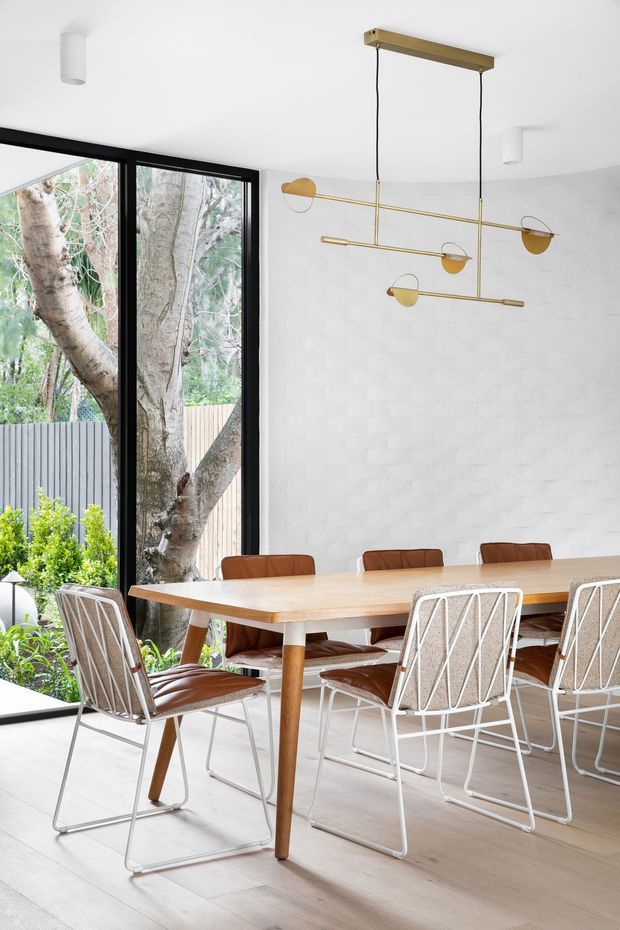 Bright contemporary dining room with a large timber dining table surrounded by mixed leather and woven chairs. Full-height black-framed glazing opens views to a leafy garden, while a sculptural brass-and-gold linear pendant light hangs over the table. The room has pale timber flooring, white walls and ceiling, and a lightly textured white feature wall.