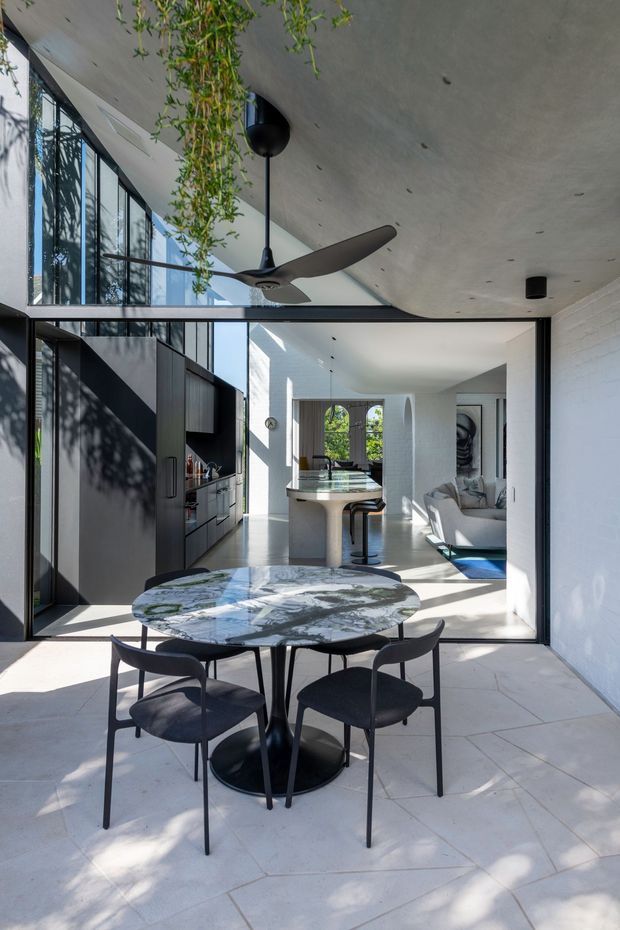 A bright, modern open-plan dining and living space viewed from a tiled terrace. The foreground features a round marble dining table with four dark upholstered chairs, while large black-framed glazed openings connect to the interior. Inside are a dark kitchen with island seating, a living area, and a second dining zone. A sculptural ceiling fan, hanging greenery, polished concrete-like ceilings, white masonry walls, and abundant natural light create a refined contemporary atmosphere.
