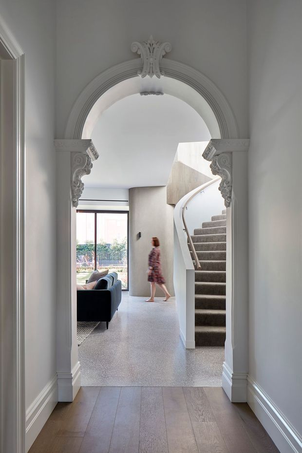View through an ornate arched opening into a bright residential entry and living area. The opening has elaborate classical plaster mouldings and carved decorative brackets, while the adjoining space features a curved staircase with carpeted treads, a pale terrazzo-like floor, a dark sofa, and large glazing to the garden. Timber flooring and detailed skirting frame the foreground hallway.