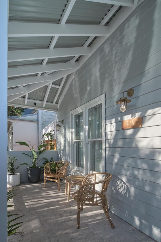 A bright covered front porch with a light blue-grey horizontal weatherboard wall, white exposed roof framing and corrugated roofing overhead, glazed entry doors, outdoor wall sconces, and a small seating area with woven rattan chairs and a table. Potted plants and a house-number plaque add residential detail.