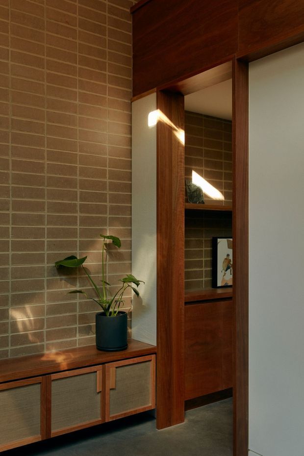 Warmly lit interior view featuring a built-in dark timber joinery wall with open shelving, a concealed doorway or passage, and a low timber cabinet with textured inset fronts. The backdrop is a full-height tan-brown brick wall, with a potted plant and framed artwork adding restrained decoration. Strong sunlight creates diagonal highlights across the timber and brick surfaces.
