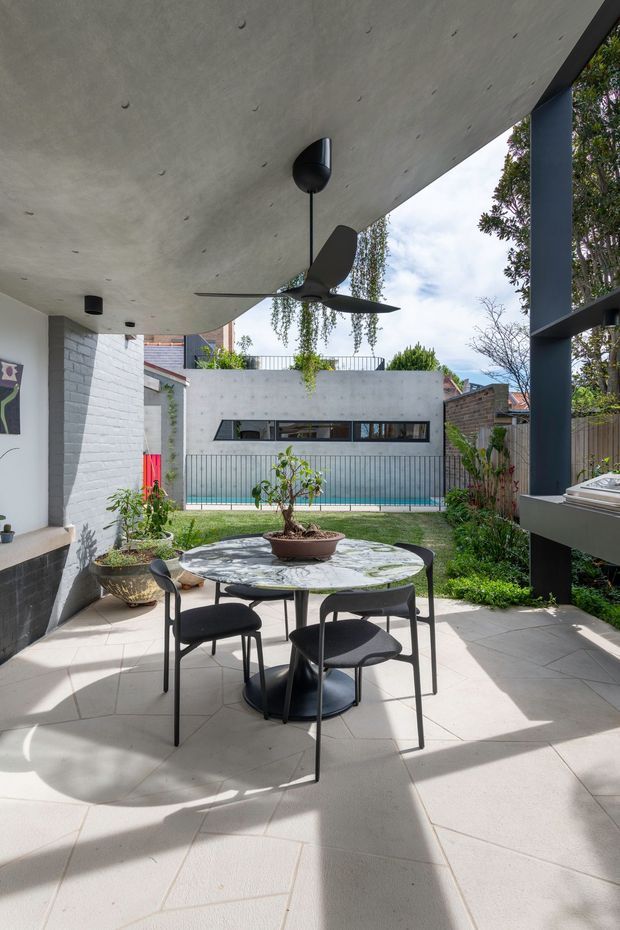 A covered outdoor dining terrace or courtyard with a round table and four dark chairs on large pale tiled paving. A ceiling fan hangs beneath a smooth concrete soffit, with dark structural posts and a built-in outdoor counter or kitchen element at right. The space opens onto a landscaped courtyard with a narrow swimming pool, rendered concrete walls, brick boundary walls, climbing greenery, and a contemporary residential setting. The image has a clean, modern architectural character with strong sunlight and contrasting shadows.