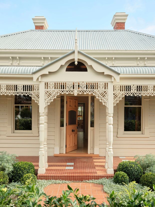 Front exterior view of a restored ornate timber house, featuring cream weatherboard cladding, a corrugated metal hipped roof, brick chimneys, decorative timber fretwork, a covered entry porch, and a terracotta tiled path and steps surrounded by manicured planting.