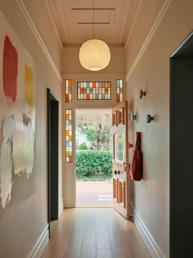 A narrow, warmly lit residential entry hall viewed toward an open timber front door. The hallway features a tiled floor, pale painted walls, ornate ceiling cornices and recessed ceiling panels, a suspended globe pendant light, stained-glass transom and sidelights around the doorway, wall sconces, and a colourful abstract artwork. The open door leads to a tiled porch and landscaped garden outside.