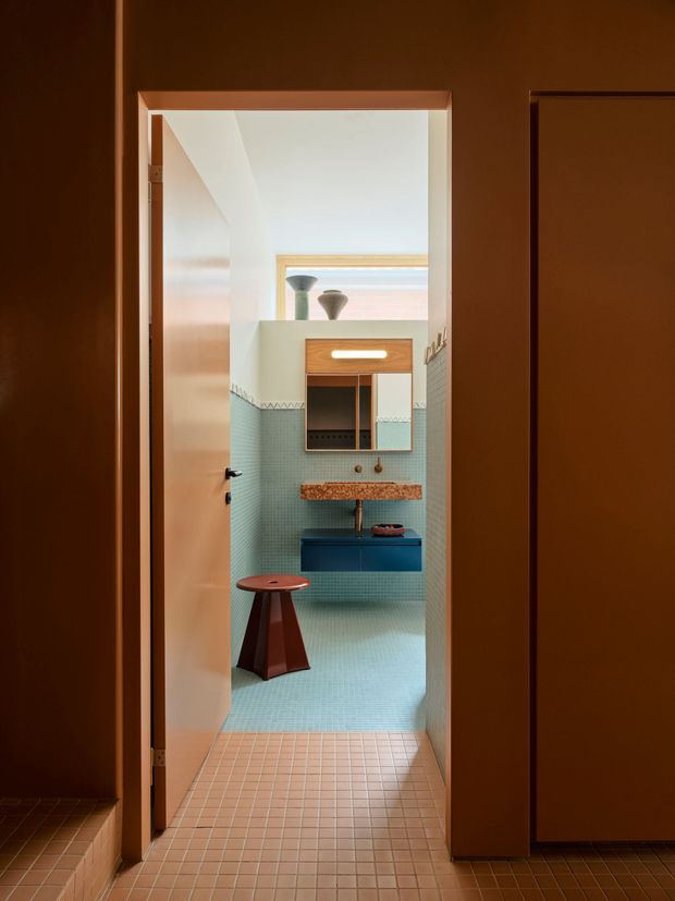 A warm, carefully framed view through an open doorway into a compact bathroom. The bathroom features pale aqua small-format tiled walls and floor, a patterned tile border, a floating deep-blue vanity, a textured terracotta-toned stone or tile benchtop, a wall-mounted mirror cabinet, and a sculptural reddish-brown stool. The surrounding hallway and door joinery are finished in rich dark brown, while daylight enters through a high rear window. The composition has a retro-inspired, refined contemporary feel with strong geometric colour blocking.