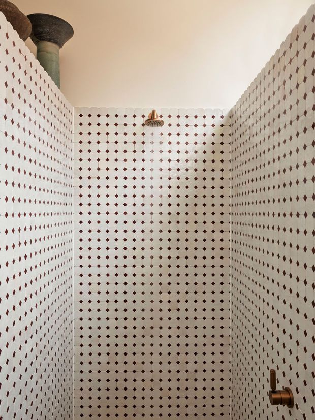 A narrow walk-in shower enclosure with three walls fully clad in repeating white ceramic tiles featuring small dark red diamond-shaped accents. A central rainfall showerhead and bronze-toned control hardware are visible. The space has a warm off-white ceiling and a decorative greenish column-like element at the upper left, creating a graphic, Moroccan-inspired bathroom interior.