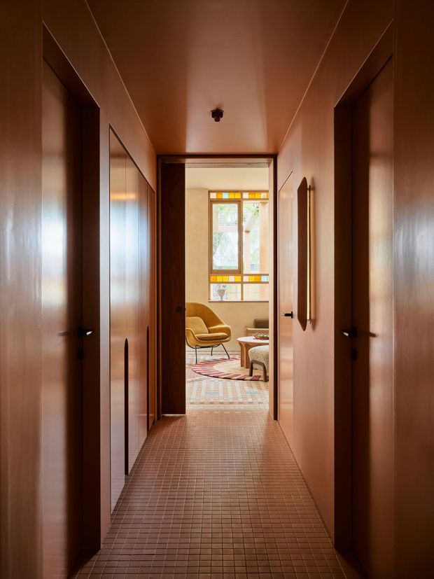 A narrow, warm-toned residential hallway lined with full-height timber veneer doors and cabinetry. The floor is finished in small terracotta-brown square tiles, and the timber-toned ceiling and walls create a continuous enveloping effect. An open doorway at the end reveals a softly furnished sitting room with a colourful geometric tiled floor, patterned rug, sculptural lounge chair, and a stained-glass-style window with amber and green detailing.