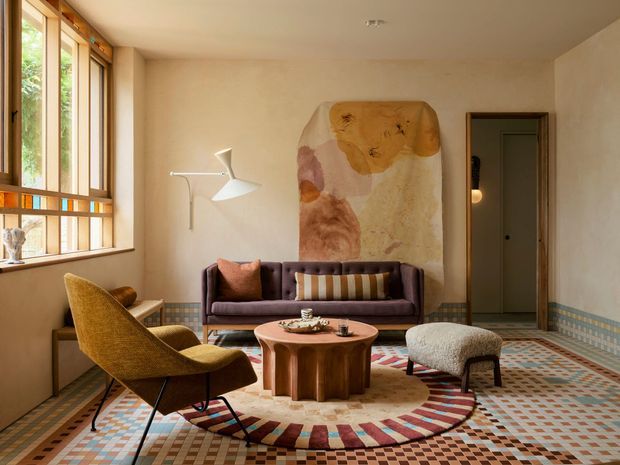 A warm, eclectic living room with a purple sofa, mustard lounge chair, sculptural round wooden coffee table, textured ottoman, and a large abstract artwork. The room features expansive timber-framed windows, softly plastered walls, a patterned geometric tiled floor and decorative tiled wall border, with layered rugs and a floor lamp creating a cosy, artistic atmosphere.