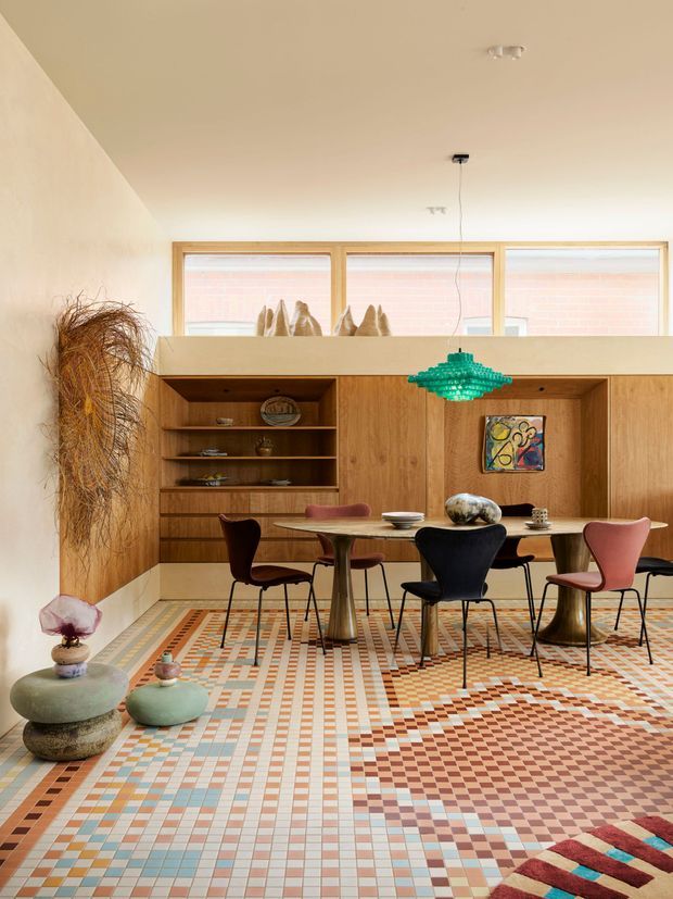 A colourful dining and living area with a long timber built-in wall unit incorporating open shelving, cabinetry and a dining nook. The room features a highly patterned geometric tiled floor in terracotta, cream, blue and grey, sculptural mid-century style dining furniture, a textured turquoise pendant light, and warm timber-framed windows. Decorative objects and artwork add an eclectic, playful character.