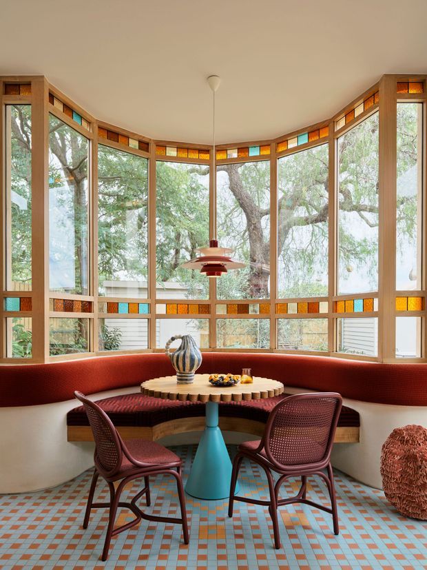 A bright, curved bay-window dining nook surrounded by expansive timber-framed glazing. The room features a built-in curved red upholstered banquette, a round timber dining table, two woven burgundy chairs, a sculptural turquoise pedestal, and a distinctive retro pendant light. Small coloured glass transoms and a patterned blue, terracotta, and cream tiled floor add a playful mid-century character, with mature trees visible outside.