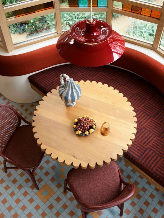 Overhead view of a vividly styled dining nook with a large scalloped-edge light timber table, curved built-in upholstered banquette, two burgundy dining chairs, and a glossy red pendant light. The space features timber-framed bay windows with patterned stained-glass transom panels, a bold geometric patterned banquette cushion, and a multicoloured checkerboard tile floor. A decorative ceramic vase, fruit bowl, and drink sit on the table.