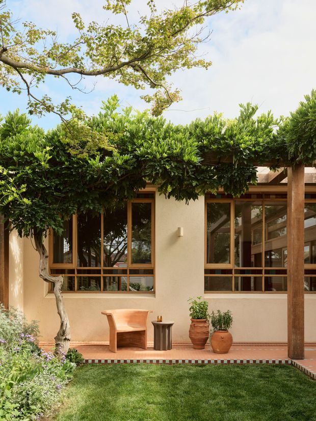 A warm, low-rise residential courtyard facade surrounded by dense greenery. The cream rendered walls feature large timber-framed windows and glazed openings, with a covered timber structure at right. A tiled terrace with decorative border leads to a manicured lawn, sculptural wooden chair, small side table, and terracotta planters. Mature trees and hedging create a leafy, private garden setting.