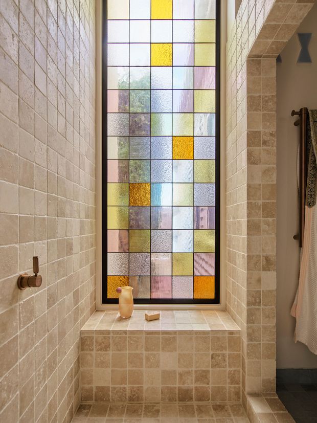 A warm, traditional bathroom with a walk-in tiled shower and a deep tiled window seat beneath a tall decorative stained-glass window. Small square beige and cream tiles cover the shower walls, ledge, seat, and lower surfaces. The multicoloured geometric stained glass provides the main feature, while a partial towel rail and hanging towels are visible at right.