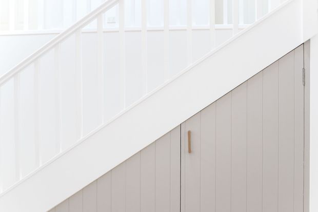 Bright minimalist interior detail showing a white stair balustrade and staircase framing above built-in vertical-panel storage beneath the stairs. The cabinetry is painted a soft light grey with a simple timber handle, while white wall panelling and abundant natural light create a clean, refined appearance.