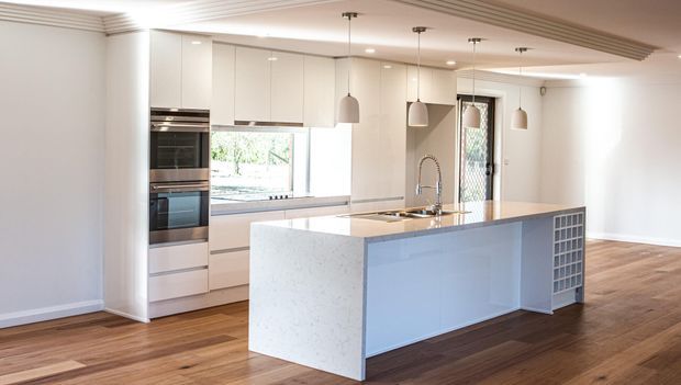 Bright contemporary kitchen with white full-height cabinetry, integrated stainless-steel appliances, a large pale stone island with waterfall end panels, pendant lighting, timber flooring, and a decorative ceiling detail. A window and glazed external door provide natural light.