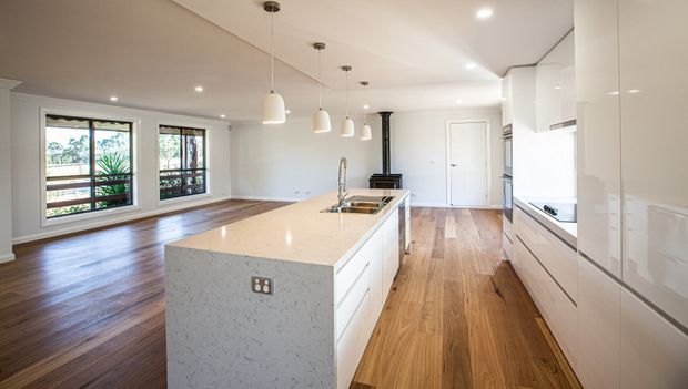 Bright open-plan kitchen with a large white stone island, integrated sink and seating potential, extensive white cabinetry, timber flooring, pendant lights, and adjoining living/dining space. Large windows provide views to greenery and a deck or balcony beyond.