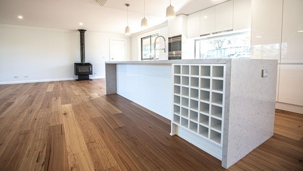 Bright open-plan kitchen and living area with extensive white flat-panel cabinetry, a large pale stone island incorporating open wine storage, timber flooring, and a freestanding black wood-burning stove. Pendant lights and broad windows add natural light to the clean, contemporary space.