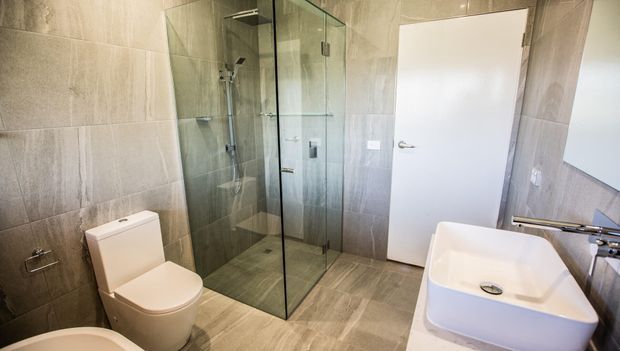 Contemporary bathroom featuring large-format grey stone-look wall and floor tiles, a frameless glass walk-in shower, white toilet, and a freestanding rectangular white vanity basin. A white hinged door and chrome fixtures complete the clean, neutral scheme.
